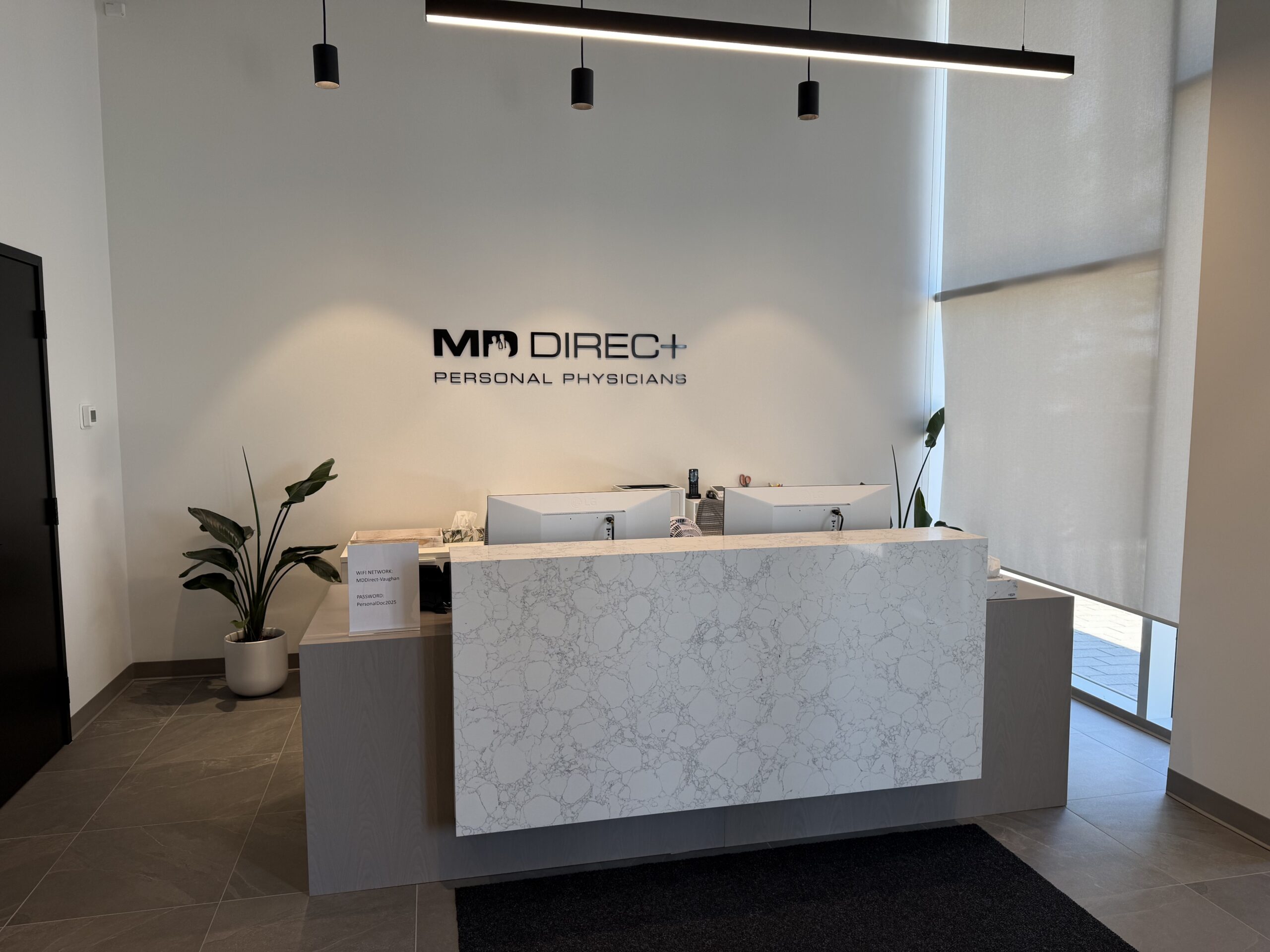 Modern medical office reception area with a marble-front desk, two computer monitors, potted plants, and a sign on the wall reading “MD DIREC+ PERSONAL PHYSICIANS.” Sunlight filters through large window blinds, highlighting a welcoming space dedicated to executive health care Toronto professionals trust.